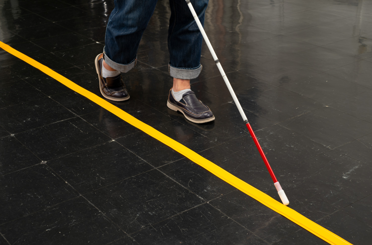 床に真っ直ぐに引かれたココテープをガイドに、白杖を持った人が歩いている。